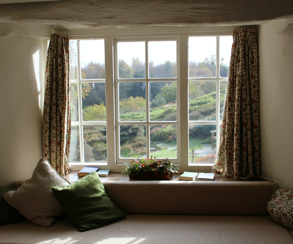 A cozy window seat with cushions and books, framed by floral curtains, overlooking a lush garden.